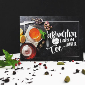 Postkarte mit der Aufschrift "Abwarten und einen im Tee haben.", sowie Teetasse, Teekanne und Kräutern vor schwarzem Hintergrund auf weißem Tisch.