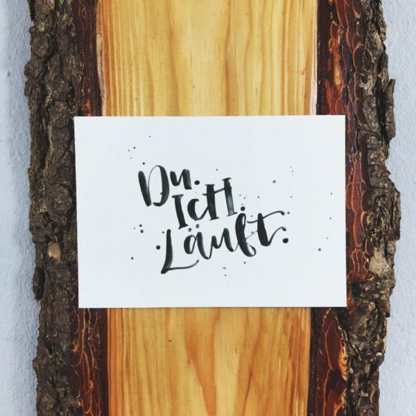 Postkarte aus Recycling Papier mit der Aufschrift "Du. Ich. Läuft." auf einer Holzplanke mit Rinde. Designt von Mint & Limes.