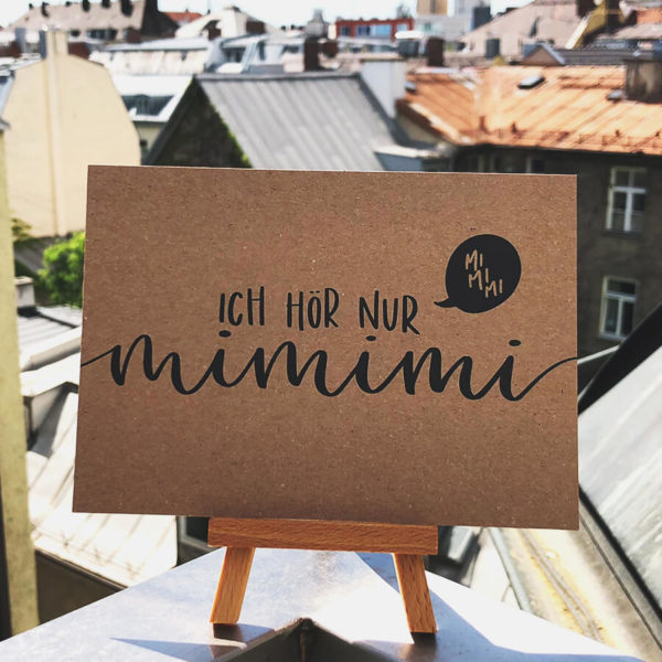 Postkarte aus Kraftkarton mit schwarzer Aufschrift "Ich hör nur mimimi" auf kleinem Holzaufsteller in der Sonne mit Hausdächern im Hintergrund. Designt von Mint & Limes.
