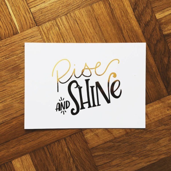 Postkarte mit der goldenen Aufschrift "Rise and shine" auf Parkettboden fotografiert.