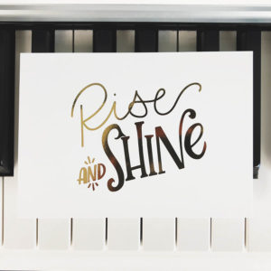 Postkarte mit der goldenen Aufschrift "Rise and shine" auf Klaviertasten liegend. Designt von Mint & Limes.
