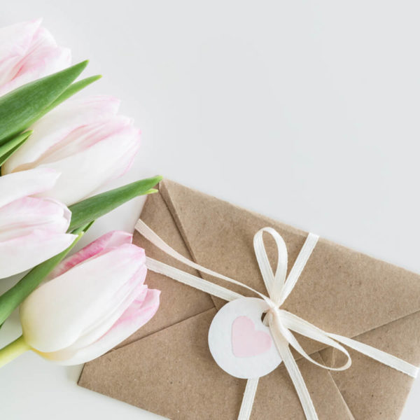 Kraftpapierumschlag mit weißem Band und Herzanhänger neben weißen Tulpen.