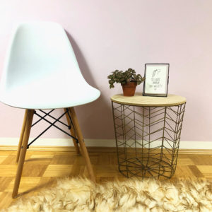 Goldener Beistelltisch aus Metall mit Holzdeckel auf dem eine Blume und ein Bilderrahmen steht. Daneben steht ein moderner weißer Stuhl vor einem Schafsfell.