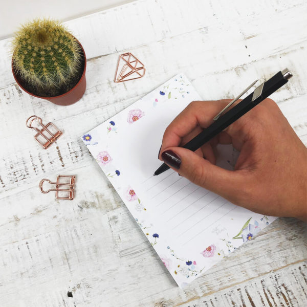 Frauenhand mit Stift, die auf einen Notizblock mit Blumenmuster schreibt. Das Bild hat einen hellen Hintergrund und ist mit Kaktus und rosé goldenen Paper Clips dekoriert.