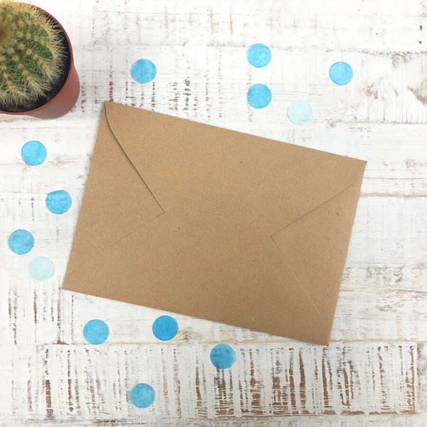 Briefumschlag aus Kraftpapier auf hellem Hintergrund und mit blauen Konfetti, sowie einem Kaktus als Dekoration.