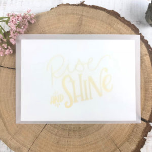 Halb transparenter Briefumschlag mit Karte "Rise and shine" auf einem aufgeschnittenen Baumstumpf neben einer Blume.