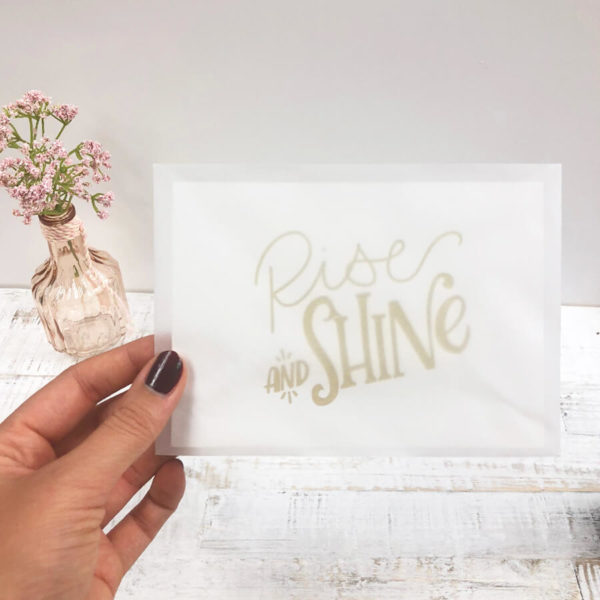Frauenhände mit Nagellack halten einen halb transparenter Briefumschlag mit Karte "Rise and shine" vor einem hellen Hintergrund.