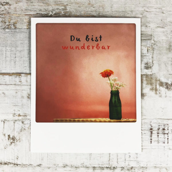 Polaroid Karte mit Aufschrift "Du bist wunderbar" und Blumenvase auf einem Tisch vor einer roten Wand.