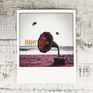 Polaroid Karte mit Aufschrift "Happy Bee-Day" und zwei Bienen, die um ein Megaphone fliegen.