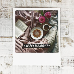Polaroid Karte mit Aufschrift "Happy Birthday" und gemütlichem Bett mit Buch, Blumen und Frühstückstablett.