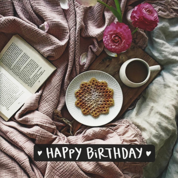 Nahaufnahme der Polaroid Karte mit Aufschrift "Happy Birthday" und gemütlichem Bett mit Buch, Blumen und Frühstückstablett.