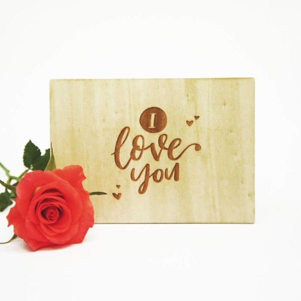 Holzpostkarte mit Aufschrift "I love you". Neben einer roten Rose vor weißem Hintergrund.