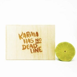 Holzpostkarte mit Aufschrift "Karma has no Deadline". Neben einer aufgeschnittenen Limette vor weißem Hintergrund.