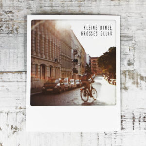 Polaroid Karte mit Aufschrift "Kleine Dinge, großes Glück" und Mann auf einem Fahrrad, der auf einer Straße fährt an deren Rand viele Autos parken.