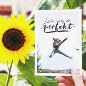 Postkarte mit Aufschrift "Lieber mutig als perfekt" und einer jungen Frau, die vor Freude in die Luft springt. Neben der Karte ist eine Sonnenblume. Designt von Mint & Limes.