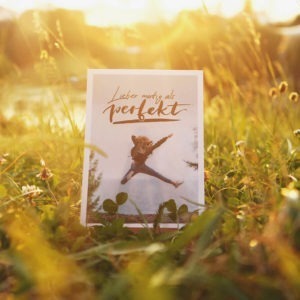 Postkarte mit Aufschrift "Lieber mutig als perfekt" und einer jungen Frau, die vor Freude in die Luft springt. Aufgenommen im Sonnenuntergang.