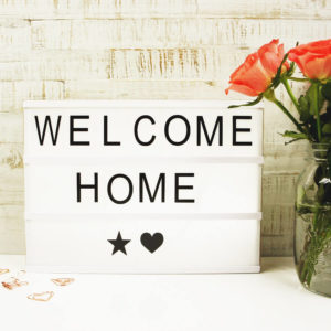 Lightbox mit Holz Gehäuse und der Aufschrift "Welcome Home" neben einer Blume und mehreren rosé goldenen Paper Clips vor hellem Hintergrund.