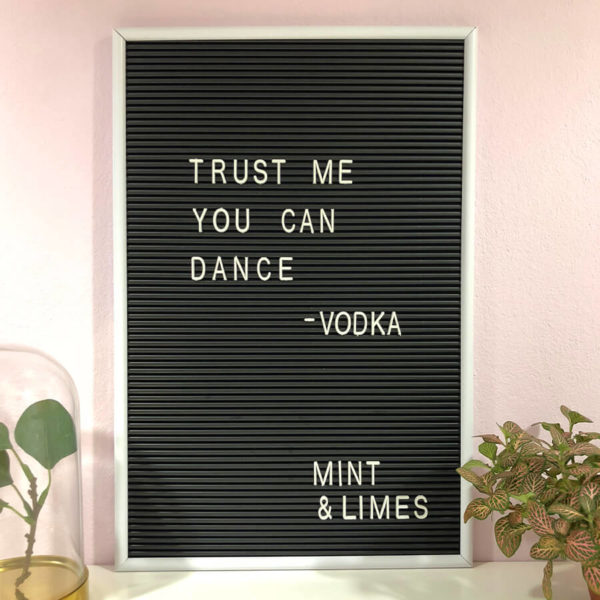 Schwarzes Message Board mit weißen Buchstaben und der Aufschrift "Trust me you can dance - Vodka" neben zwei Blumen als Dekoration.