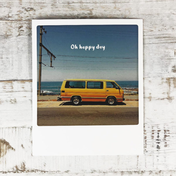 Polaroid Karte mit Aufschrift "Oh happy day" und gelbem Kleinbus, der am Meer parkt.