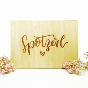 Holzpostkarte mit Aufschrift "Spotzerl" und kleinem Herz neben zwei Blumen vor weißem Hintergrund.