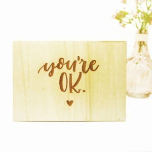 Holzpostkarte mit Aufschrift "You're OK" und kleinem Herz neben einer Vase mit einer Blume.